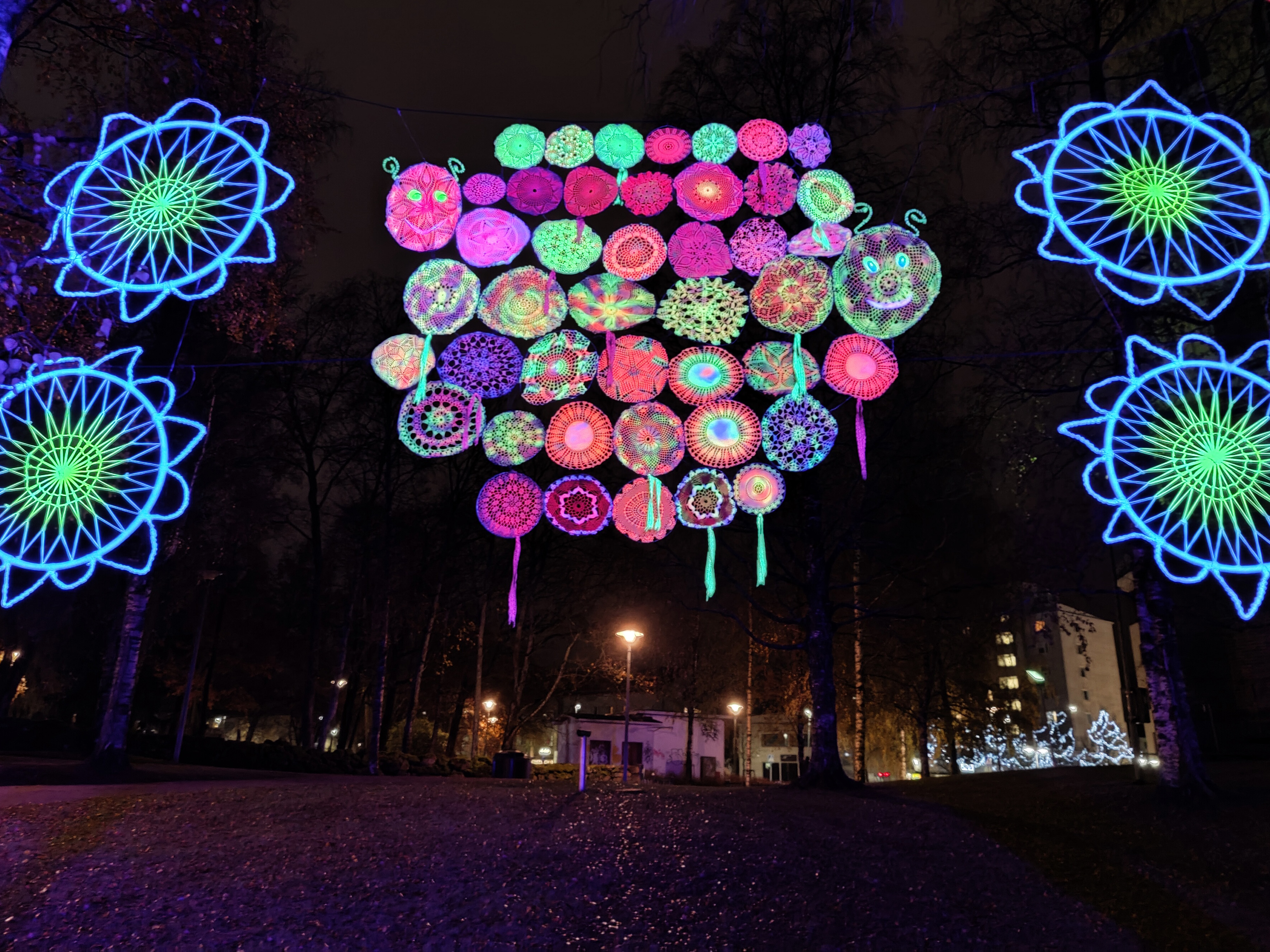 Värikkäät toukka- ja kukka-valotaideteokset ripustettuna puuhun.
